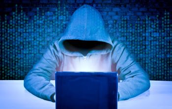 Person wearing blue sweatshirt with hood covering face to hide identity, sitting at a laptop and stealing sensitive data.